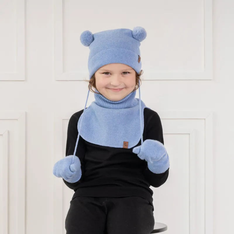 A young child with light brown curly hair and a subtle smile, is looking directly at the viewer. They are wearing a light blue knit hat with two pom-poms and earflaps that tie under the chin, matching light blue mittens, and a light blue knit neck warmer, over a black long-sleeved shirt. The background is a white paneled wall.