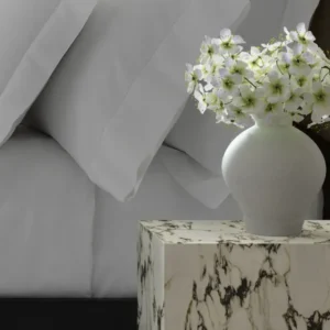 A close-up of ivory pillows on a bed beside a beige marble side table holding a white vase filled with delicate flowers. Featuring Gatsby Hemstitch bedding by Matouk. Available at Linen Society.