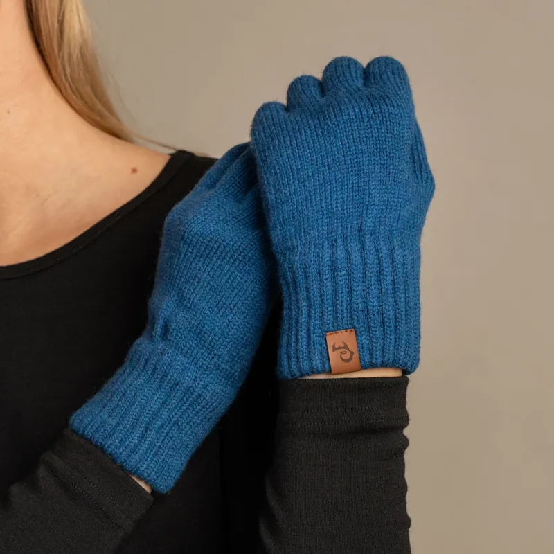 A close-up shot of a person wearing menique brand vibrant blue, ribbed knit gloves. The person is wearing a dark long-sleeved top, with the right hand holding the left wrist, showcasing the gloves' fit and the small, rectangular tan leather menique tag on the cuff of the left glove. The background is a solid, neutral gray.
