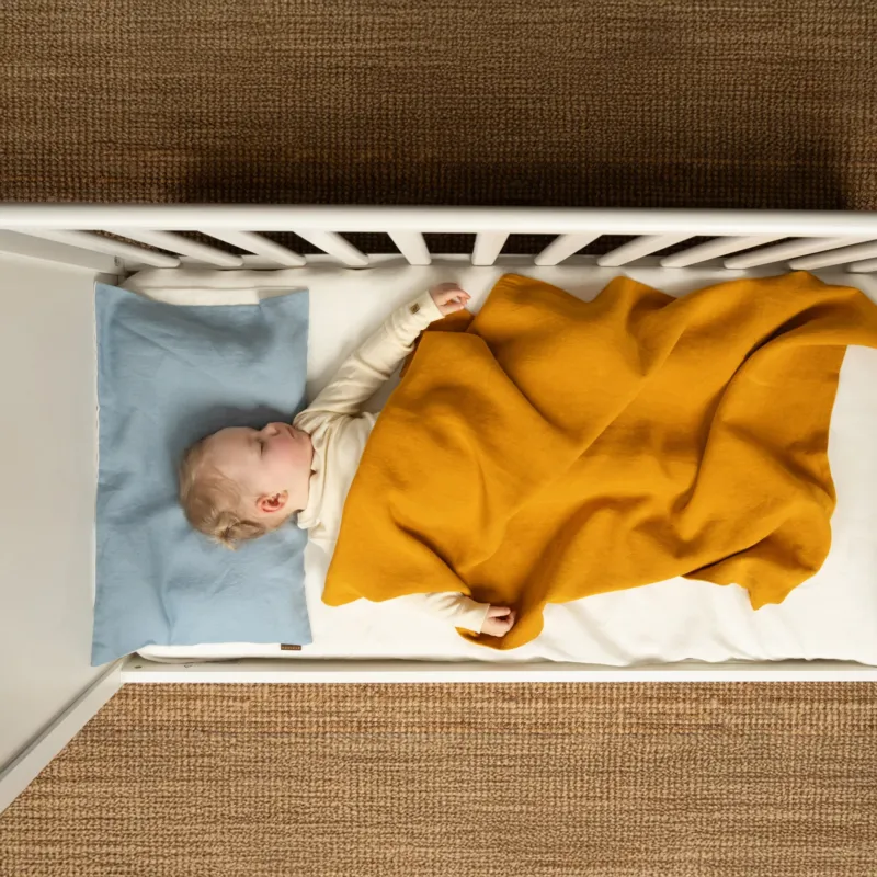 Baby sleeping in a white crib under a mustard yellow linen blanket, on a natural brown woven rug.