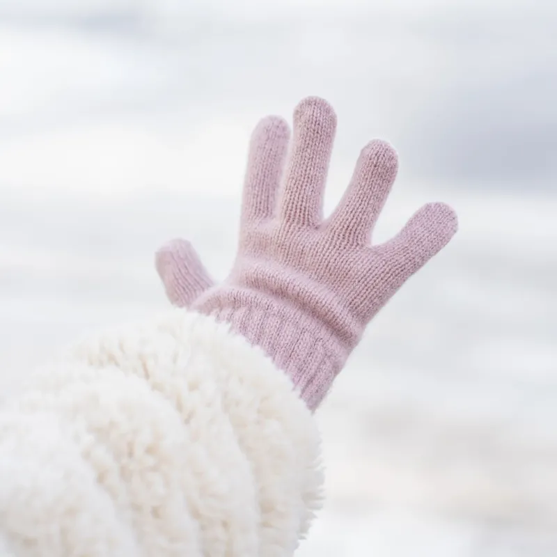 A hand wearing a light pink knitted glove with a ribbed wrist extends upwards and slightly to the left, revealing a plush, cream-colored sleeve on the arm, against a blurred light background.