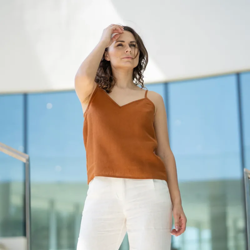A woman with brown hair wears a almond brown V-neck camisole top and white trousers, posing indoors in front of large glass windows.