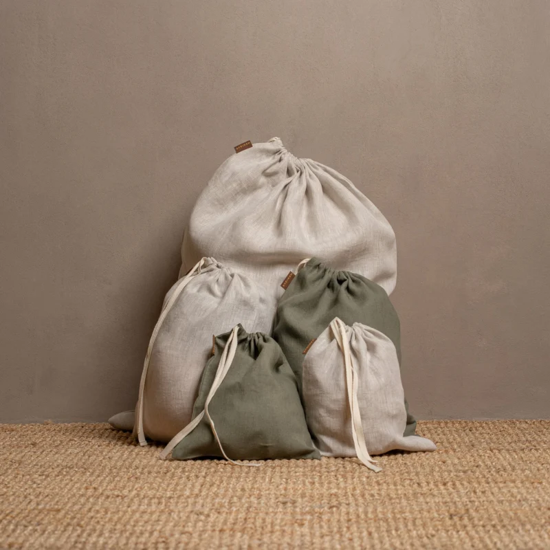 Five closed linen drawstring bags in varying sizes and colors of natural and stone green are arranged on a textured jute rug against a warm brown wall.
