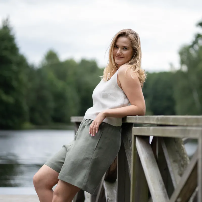 A barefoot woman strolls down a sun-dappled dirt path through a lush green forest. She is wearing a natural-colored linen crop top paired with comfortable stone green linen Bermuda shorts.