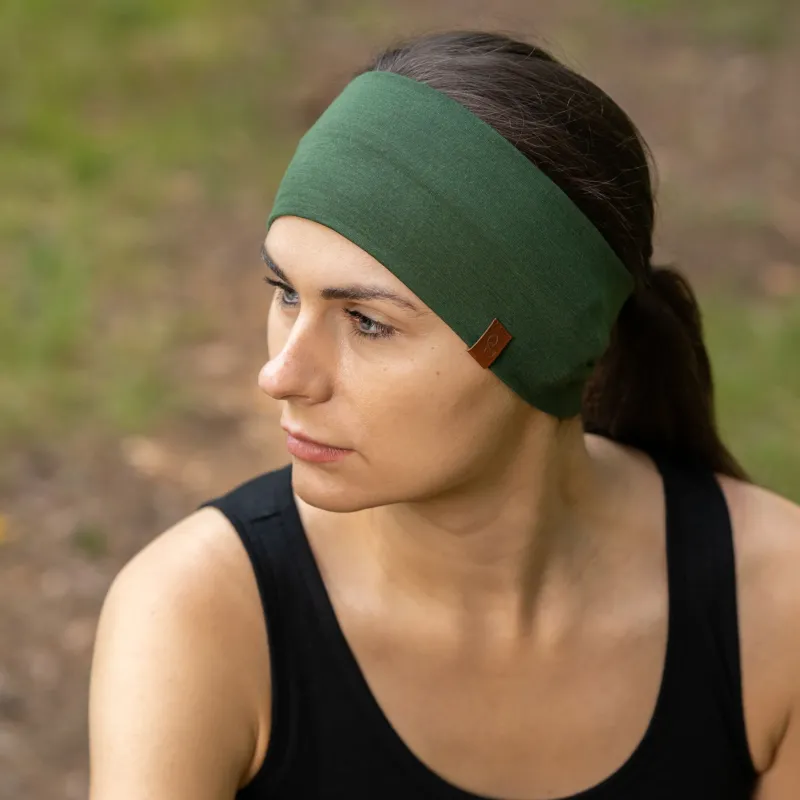 The picture features a woman sitting down in a forest, wearing the unisex dark green menique merino HEADBAND. She is also wearing a sleeveless black merino top. She is looking to the side.