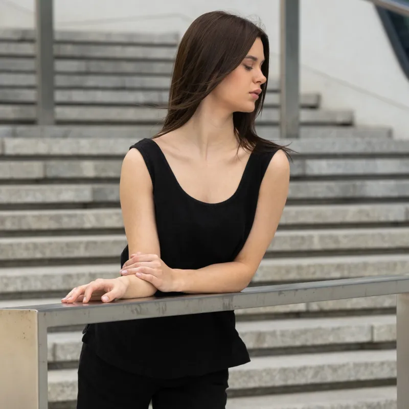A young girl posing outdoors in a city on a concrete stairs, wearing linen pure black tank top Hana, and pure black pants dakota.