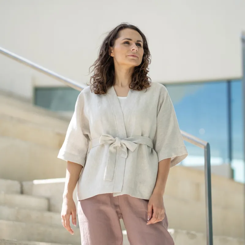 A woman with brown hair, wearing a light beige kimono-style jacket over mauve trousers, stands on indoor concrete stairs.