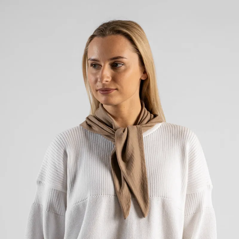 A studio portrait of a woman looking off to the left side of the frame. She is wearing a small, light beige bandana tied loosely around her neck like a scarf. She is wearing a textured white sweater, and the background is a solid light grey.