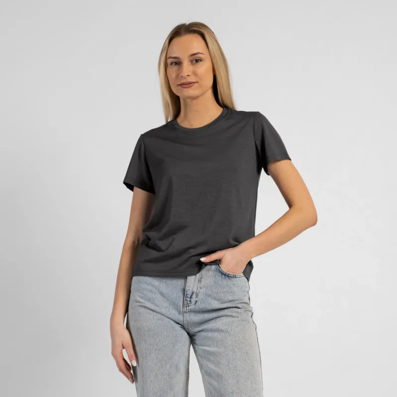 A studio portrait of a blonde woman standing and looking directly at the camera, with her left hand casually tucked into the front pocket of her light-wash blue jeans. She is wearing a regular fit, charcoal grey short-sleeved crew-neck t-shirt against a solid light grey background.