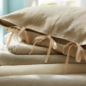 SDH Camden linen-textured pillow in natural with champagne ribbon ties, stacked with folded bedding.