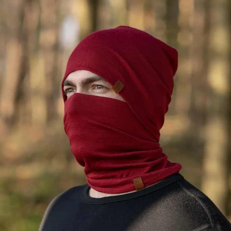 A man, standing outside in a forest during fall season, wearing the Menique Men's Merino Beanie and neck Gaiter Set in the color Royal Cherry.