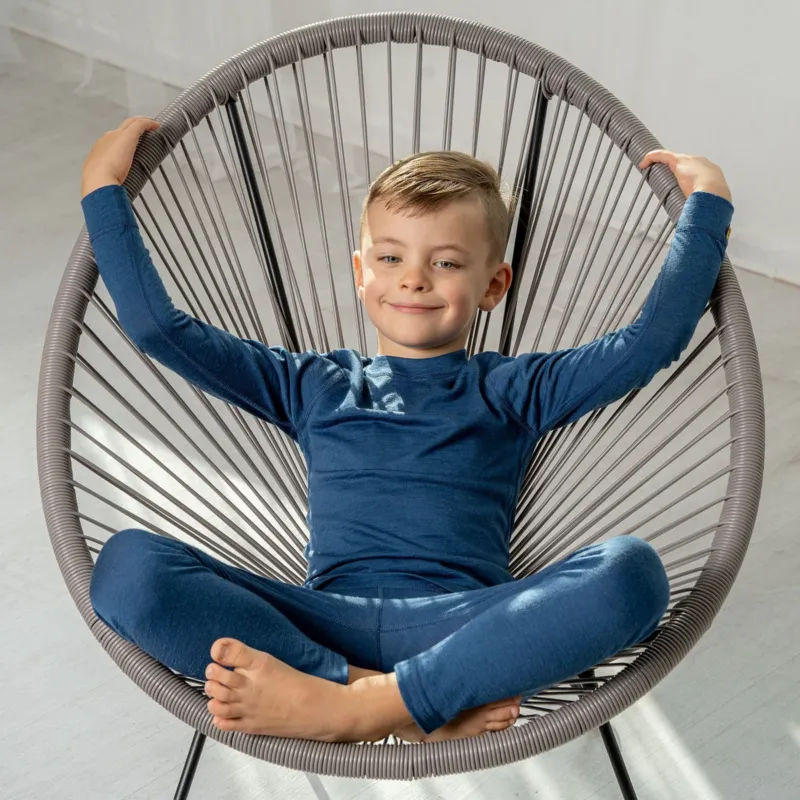 A boy sitting in a lounge chair, wearing the menique Kids' Merino 160 Long Sleeve Set in Denim color.